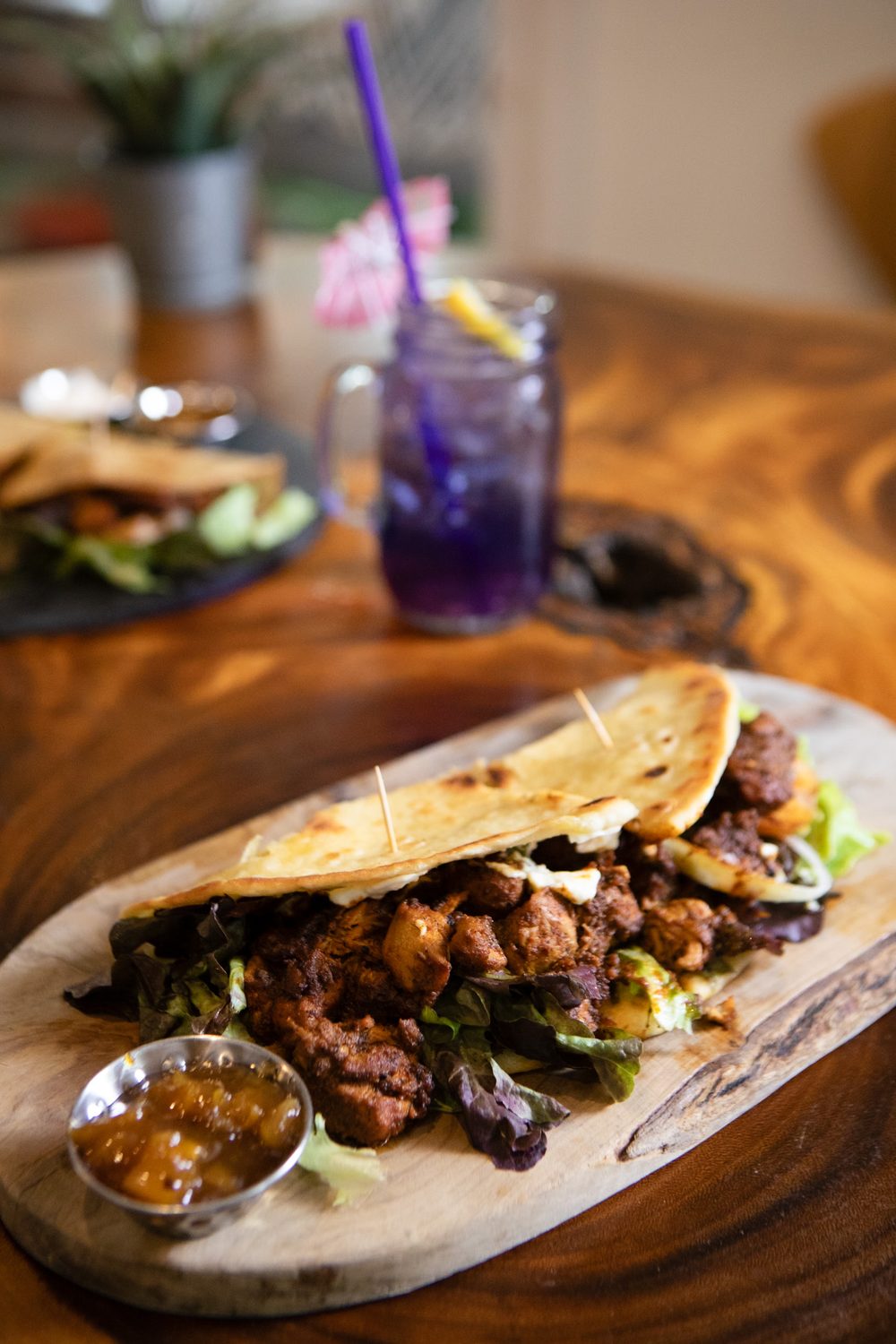 A grilled flatbread filled with jerk chicken, lettuce and tomato, served on a wooden board with a purple iced drink.