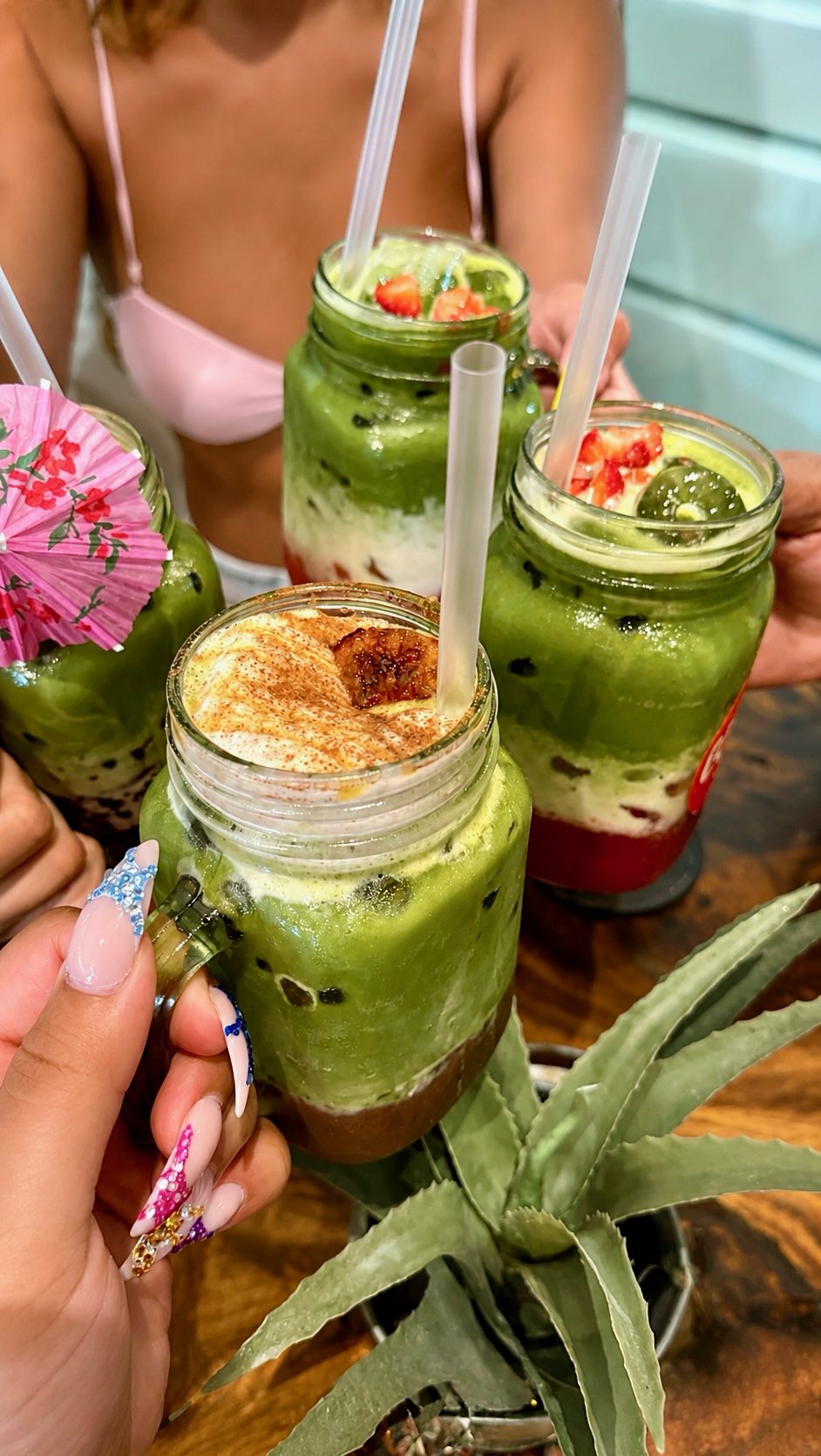 Five mason jars of iced matcha drinks on a table, with strawberry and a paper umbrella.
